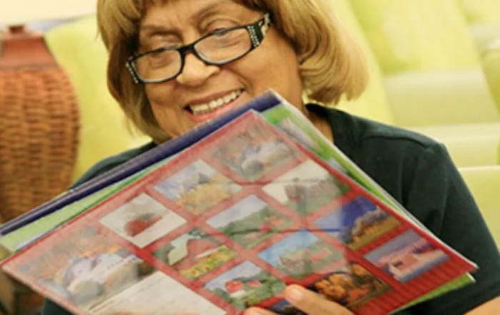 An elderly woman wearing glasses is smiling while reading a book indoors.