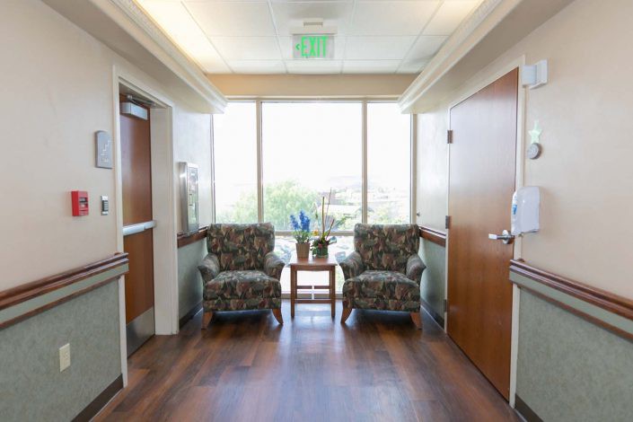 A well-lit waiting area with two patterned chairs and a small table by a large window.