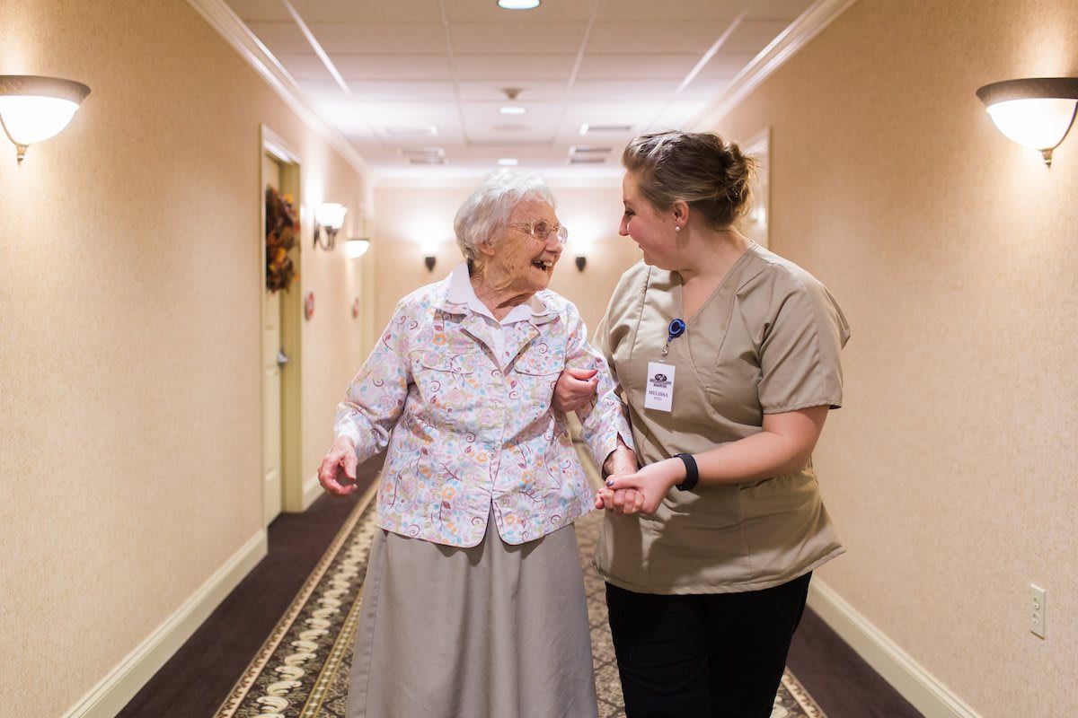A caregiver assists an elderly woman as they walk together down a hallway.