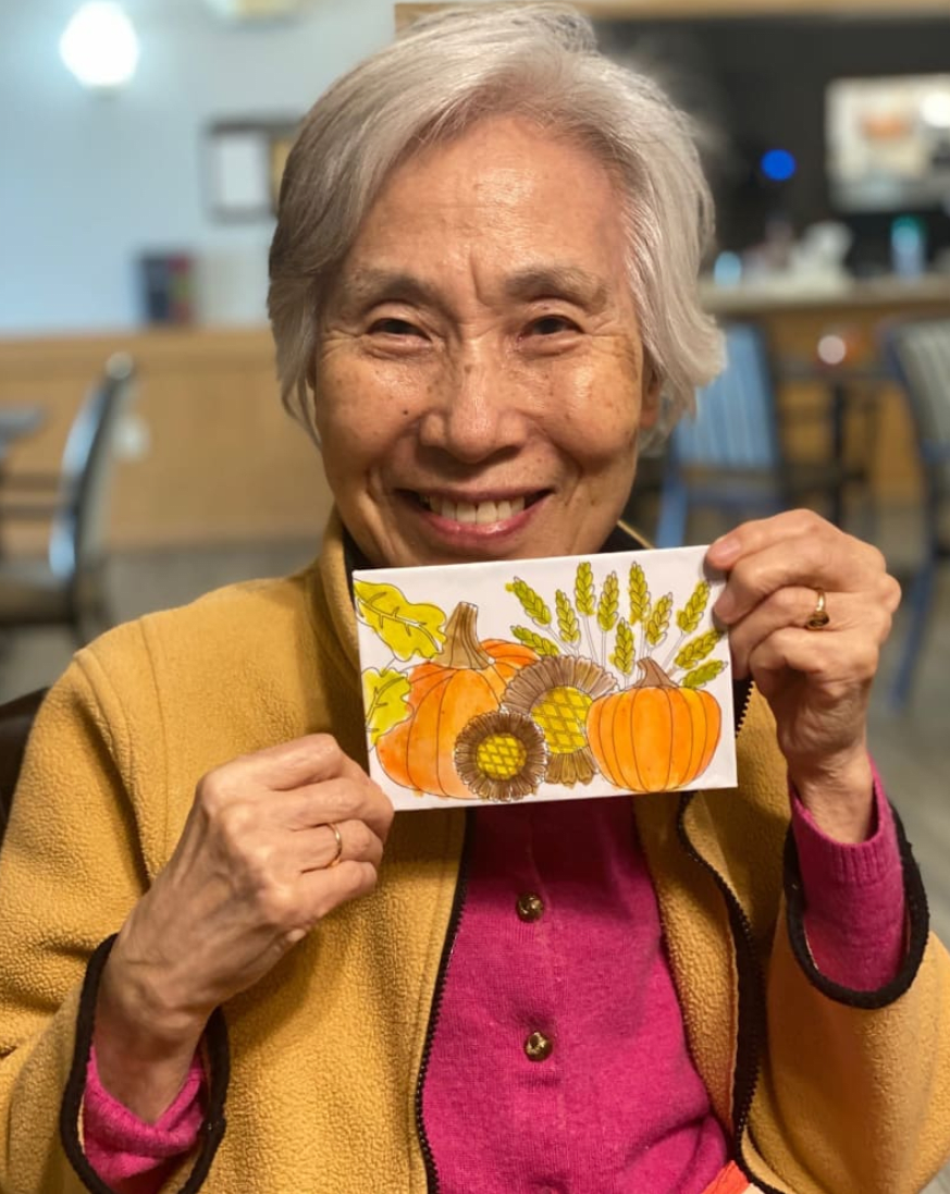 An elderly woman smiles while holding a card with an autumn-themed design.