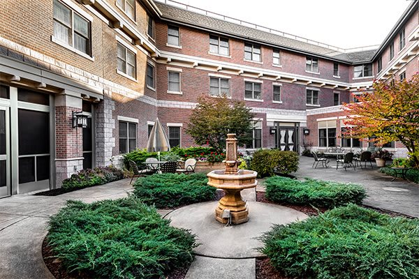A charming courtyard featuring a central fountain surrounded by lush greenery and outdoor seating, set against a brick building backdrop.
