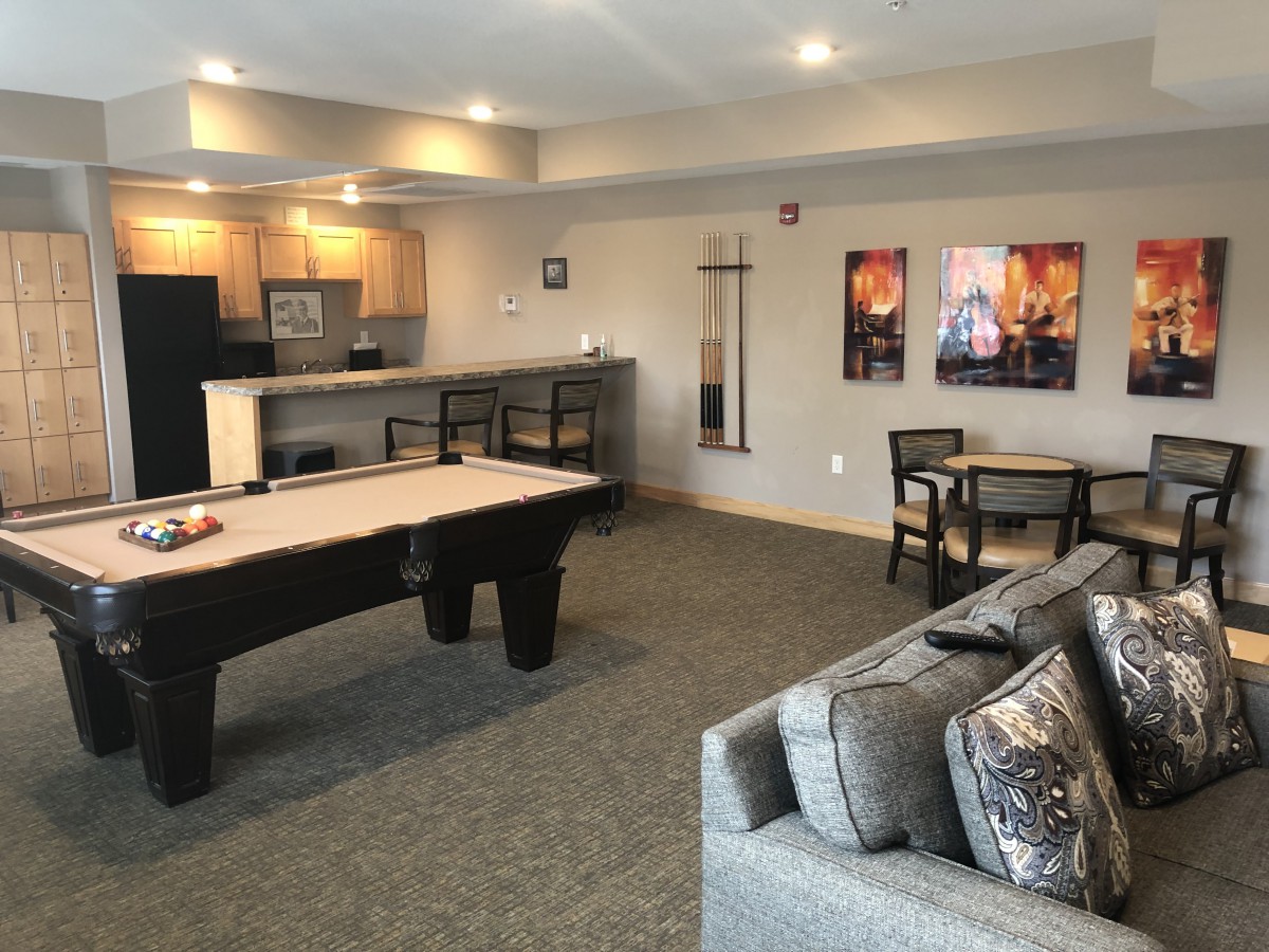 A modern game room featuring a shuffleboard table, kitchenette, and comfortable seating.