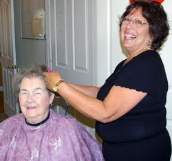 A hairstylist is giving an elderly woman a haircut, both smiling indoors.