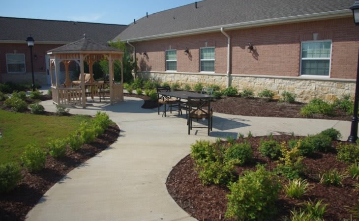 A landscaped outdoor area featuring a patio with seating, a pergola, and a walkway surrounded by shrubs and mulch in front of a building.