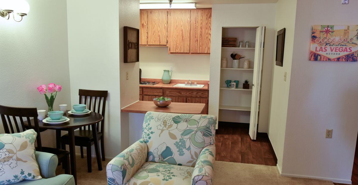 A cozy apartment interior featuring a small kitchenette with wooden cabinets and an open shelf beside a patterned armchair.