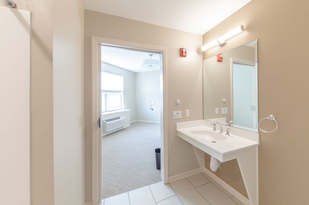 An empty bathroom with a sink and mirror, leading into a bright room.