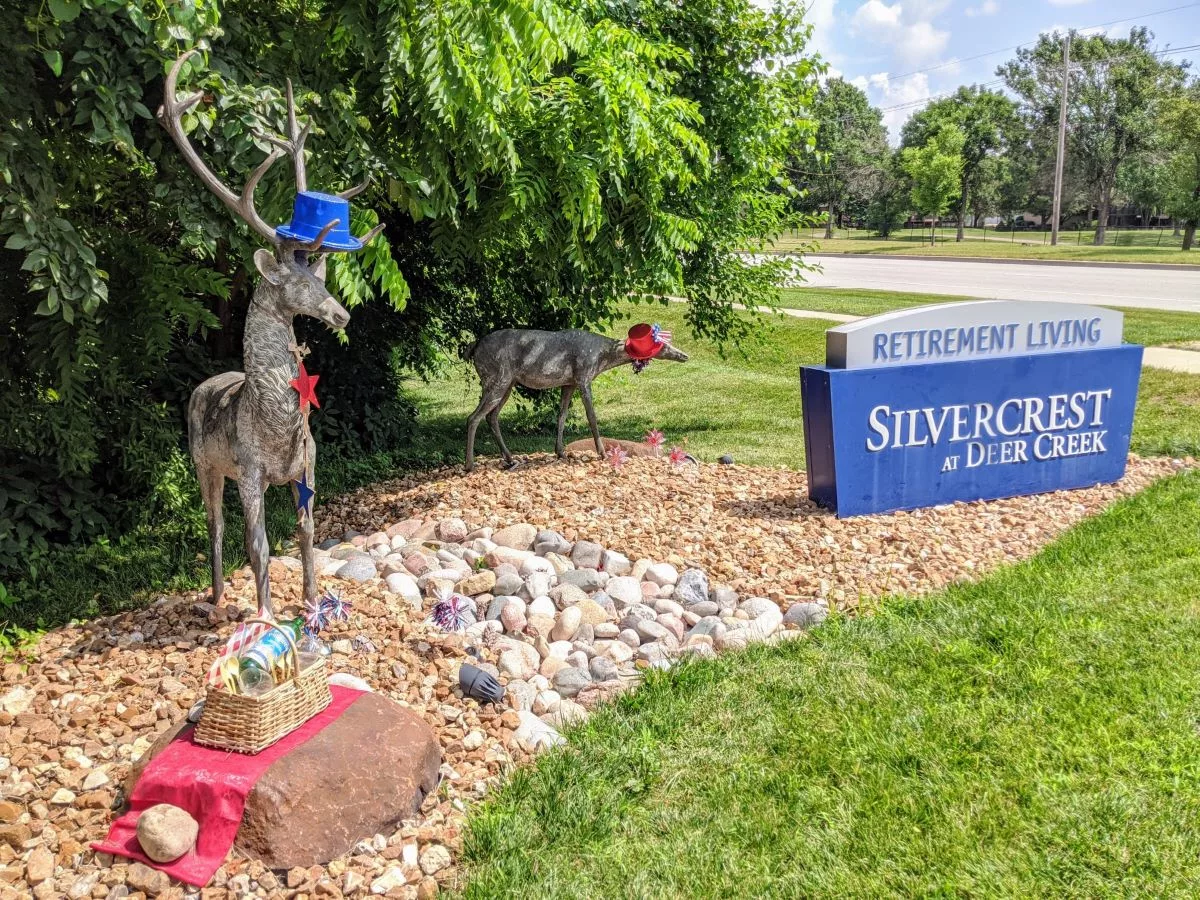 Metal deer sculptures wearing hats and ties are displayed near a sign for Silvercone at Lakewood amidst landscaped stones and greenery.
