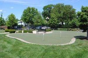 A putting green surrounded by lush trees and benches on a sunny day.