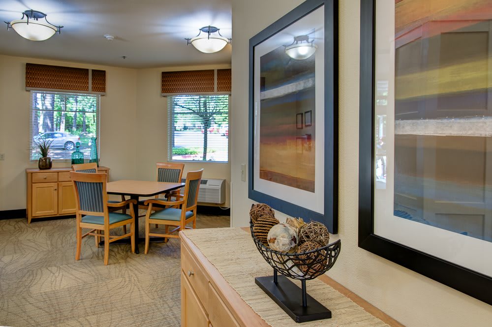A cozy living area with a wooden table and chairs, framed artwork on the wall, and natural light streaming in from the windows.