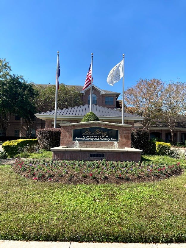 A building entrance with a prominent sign and three flags, surrounded by landscaping under a clear blue sky.