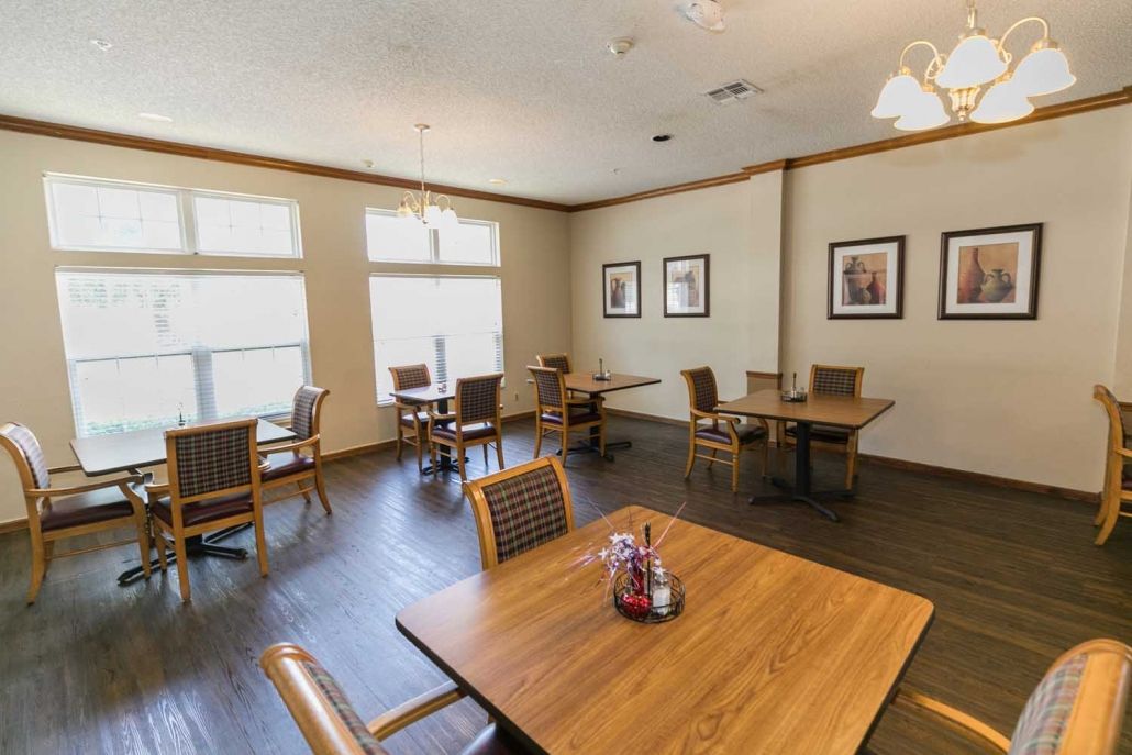 A cozy dining room with several tables and chairs, adorned with art on the walls and well-lit by large windows.