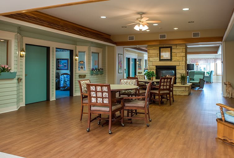A cozy interior lounge with tables and chairs arranged around a fireplace, featuring wooden flooring and a ceiling fan.