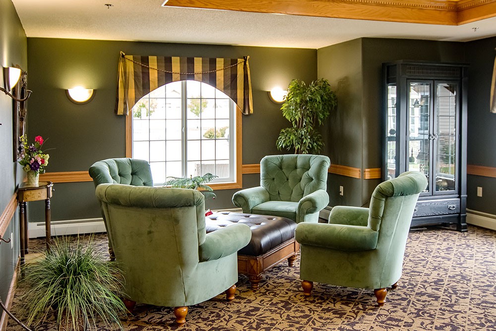 A cozy living room with green chairs arranged around a central coffee table, illuminated by natural light from a large window.