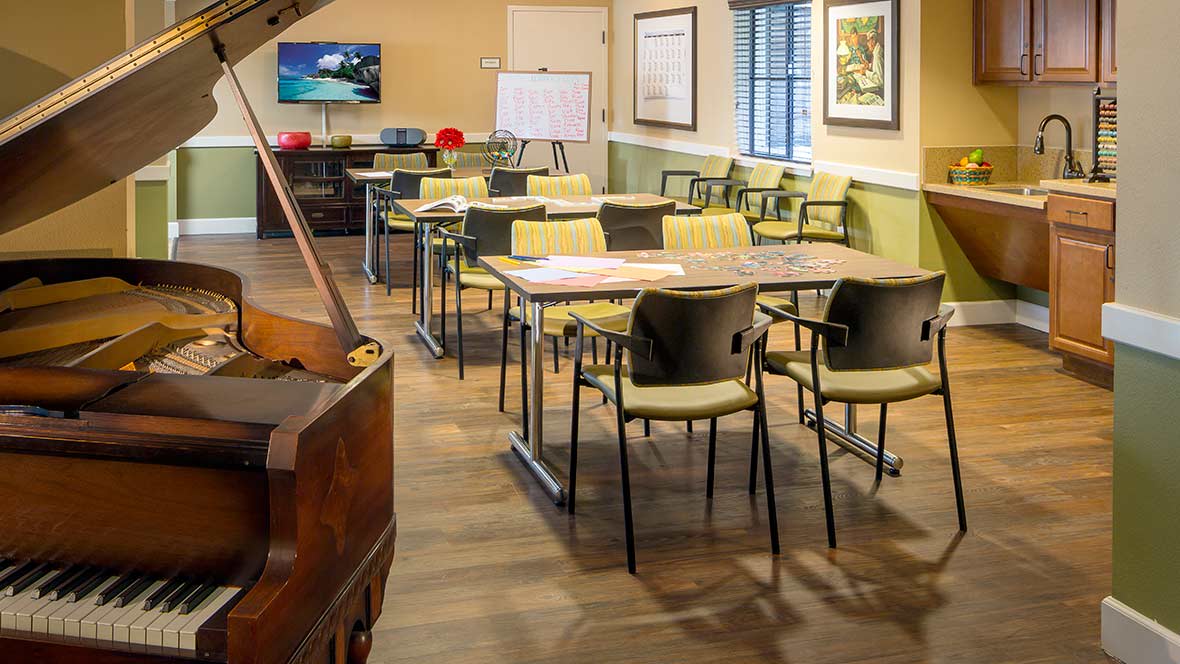 An empty classroom set up with tables and chairs, a piano in the corner, and a whiteboard at the front.