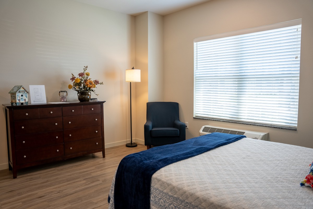 A cozy bedroom with a neatly made bed, wooden dresser, and an armchair by the window.