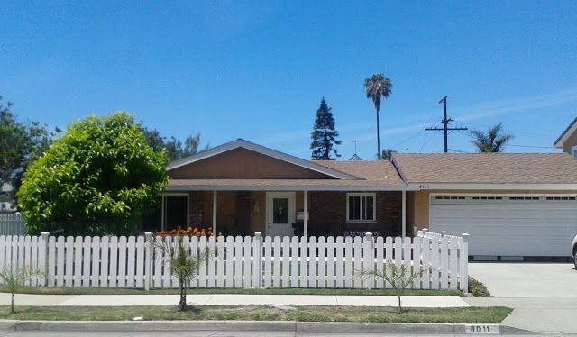 A suburban house with a white picket fence and palm trees under a clear blue sky.