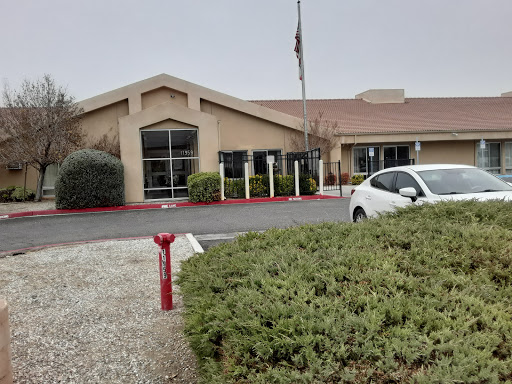A view of a single-story building with a parking lot and greenery in the foreground, set against a cloudy sky.