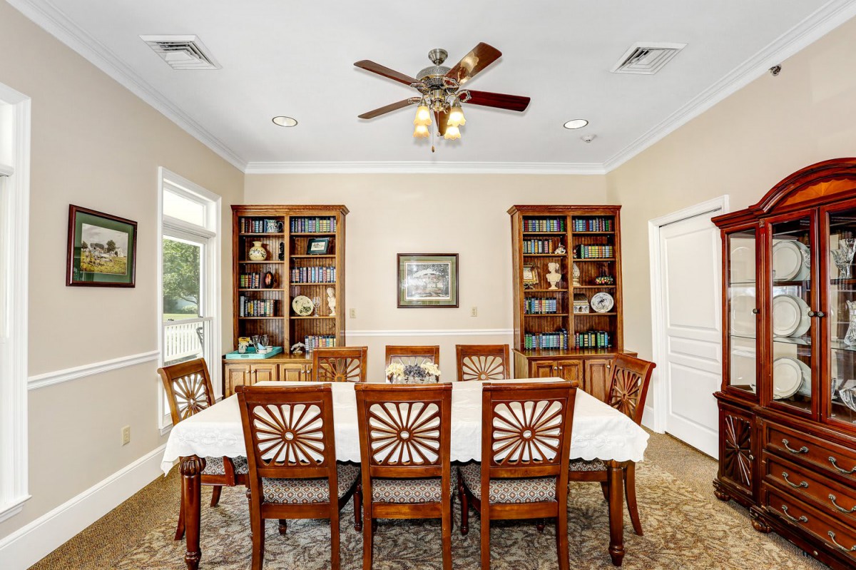 A well-furnished dining room with a large table surrounded by chairs, flanked by bookshelves and lit by a ceiling fan.