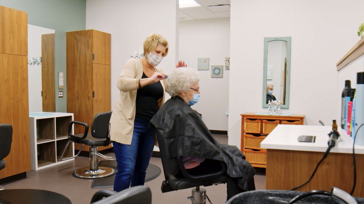 A hairstylist wearing a mask provides a haircut to an elderly client in a salon.