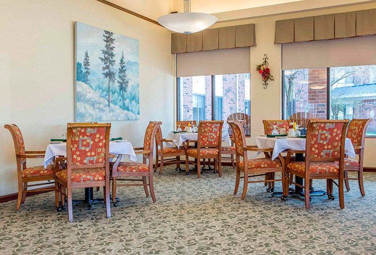 A cozy dining room with elegantly upholstered chairs and tables, featuring artwork and natural lighting from large windows.