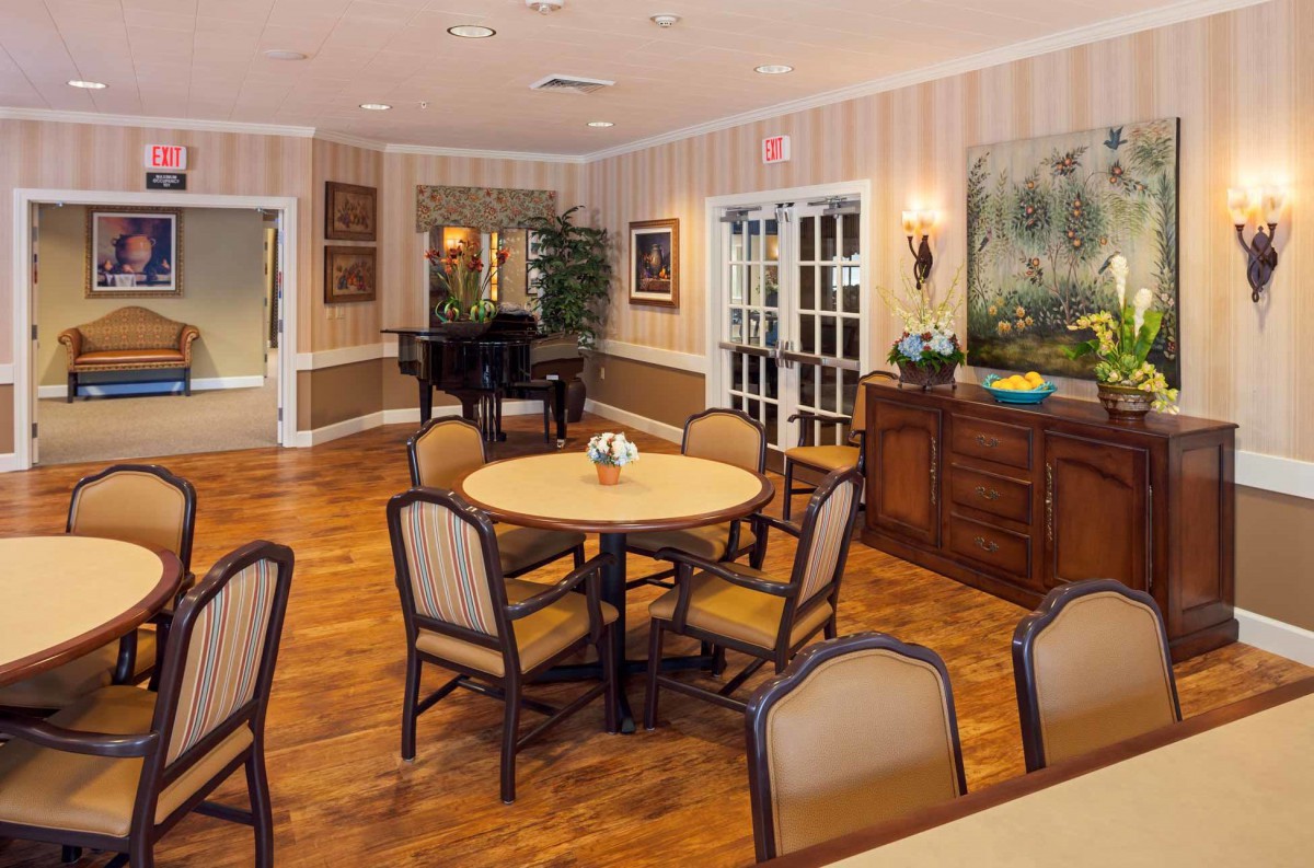 A cozy dining area with round tables and cushioned chairs on a wooden floor, featuring wall art and a piano in the background.