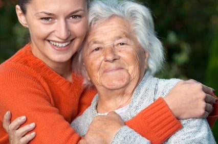 A young woman embraces an elderly woman, both smiling warmly outdoors.