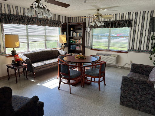 A cozy living room featuring a comfortable sofa, a dining table with chairs, and a bookshelf, illuminated by natural light.