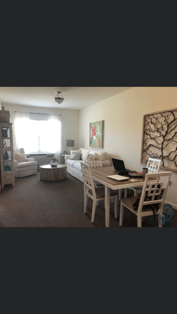 A cozy living room with a dining table and laptop setup, featuring neutral-toned furniture and natural lighting.