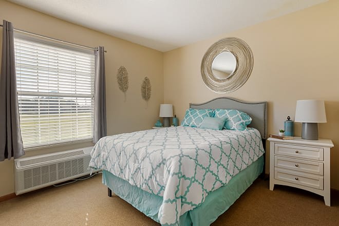 A cozy bedroom with a bed featuring patterned bedding, a lamp on the nightstand, and a circular mirror on the wall.