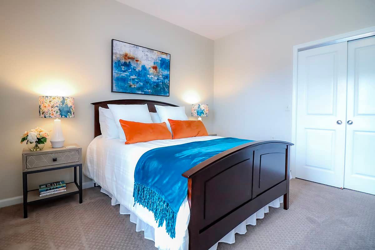 A neatly organized bedroom featuring a wooden bed with colorful pillows and a blue throw blanket, complemented by wall art and bedside lamps.