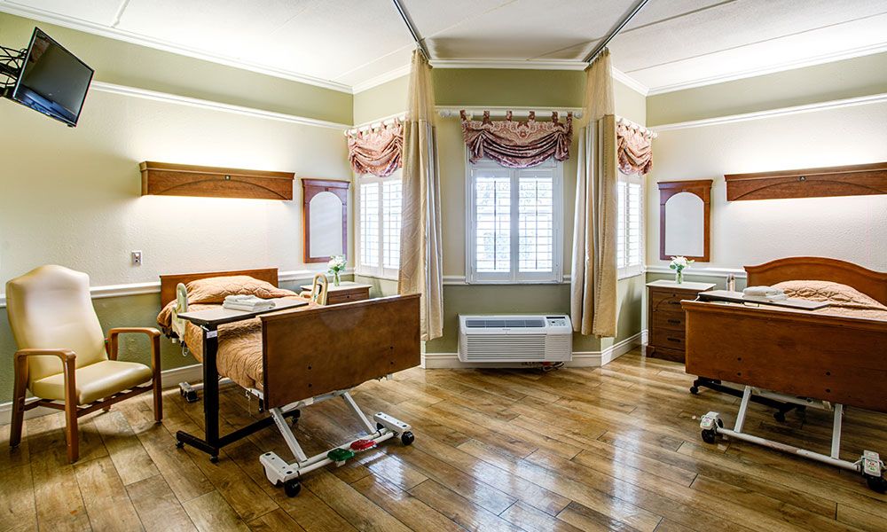 A clean and spacious hospital room with two patient beds, separated by curtains, featuring large windows for natural light.
