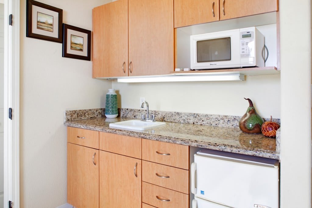 A small kitchen area with wooden cabinets, a granite countertop, a microwave, and a mini fridge.