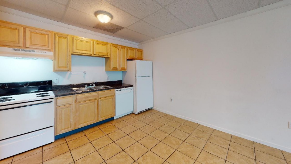 An unfurnished apartment kitchen with wooden cabinets, a refrigerator, and a tiled floor.