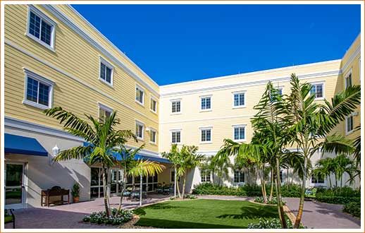 A bright courtyard featuring palm trees and a well-maintained garden, surrounded by a yellow multi-story building under a clear blue sky.