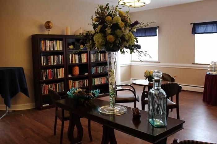 A cozy library corner with bookshelves, a large vase of flowers on a table, and chairs arranged for reading.