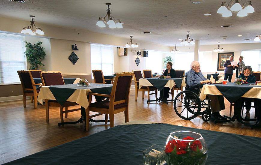 A quiet dining area in a nursing home with tables set for meals, featuring an elderly person in a wheelchair.