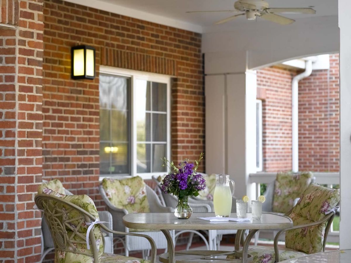 A cozy porch with wicker furniture, a table set with lemonade and flowers, against a brick wall backdrop.