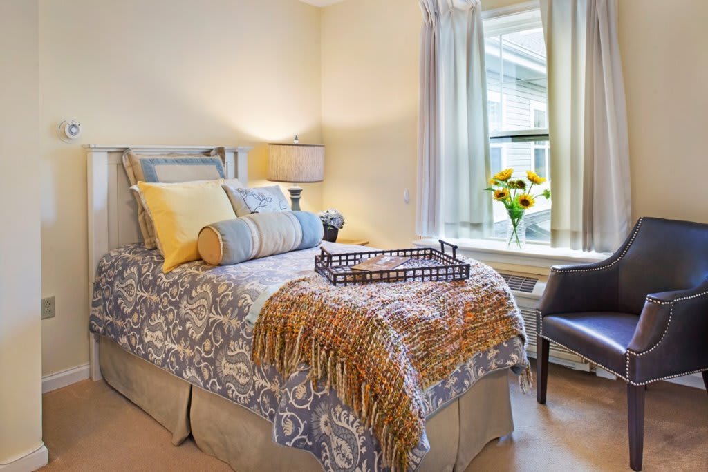 A cozy bedroom with an elegantly decorated bed, featuring patterned bedding and an assortment of pillows. A lamp sits on the bedside table, while a chair and flowers by the window add to the inviting atmosphere.