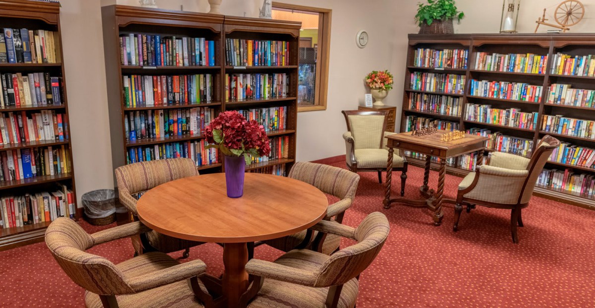 A cozy library room with bookshelves, a round table, chairs, and a vase of flowers.