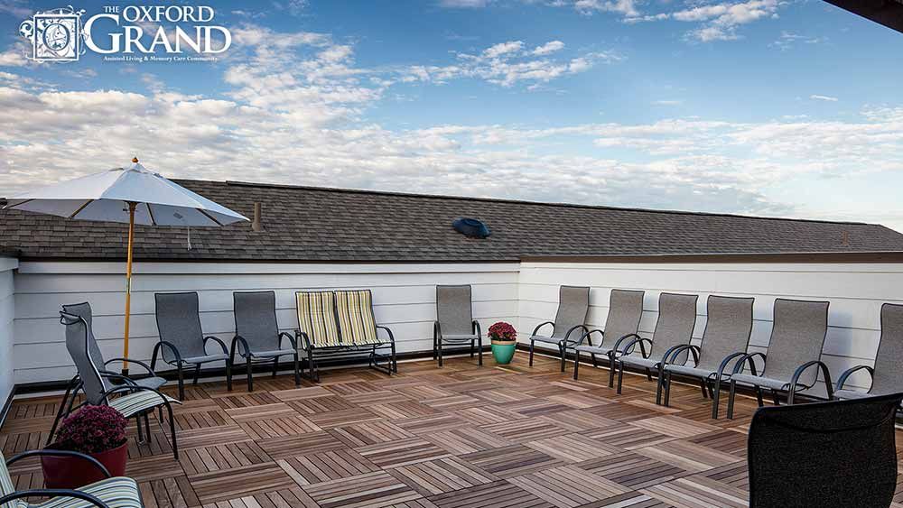 A rooftop patio with several chairs arranged on a wooden deck under a blue sky with clouds.