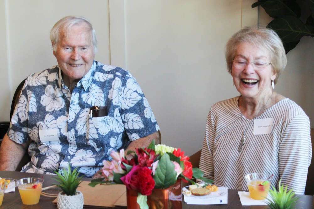 An elderly couple smiling and enjoying a celebration indoors with decorations around them.