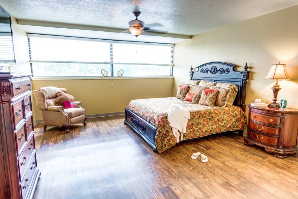 A spacious bedroom with a large bed, an armchair, and a ceiling fan, featuring a wooden floor and a wide window.