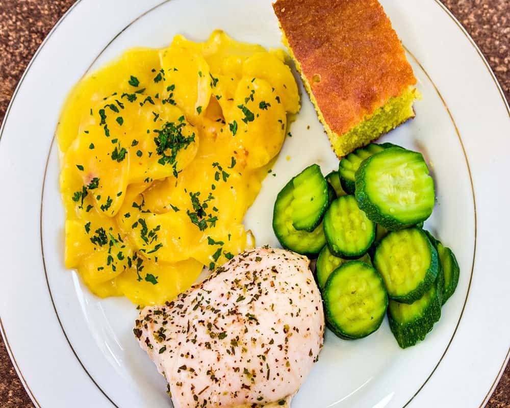 A meal consisting of seasoned chicken, sliced potatoes with herbs, cucumber slices, and a piece of cornbread on a plate.