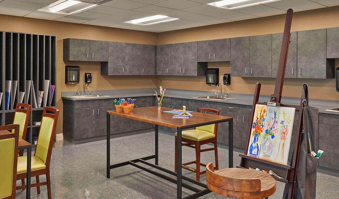 An art classroom featuring a large table with paint supplies, surrounded by cabinets and an easel with artwork.