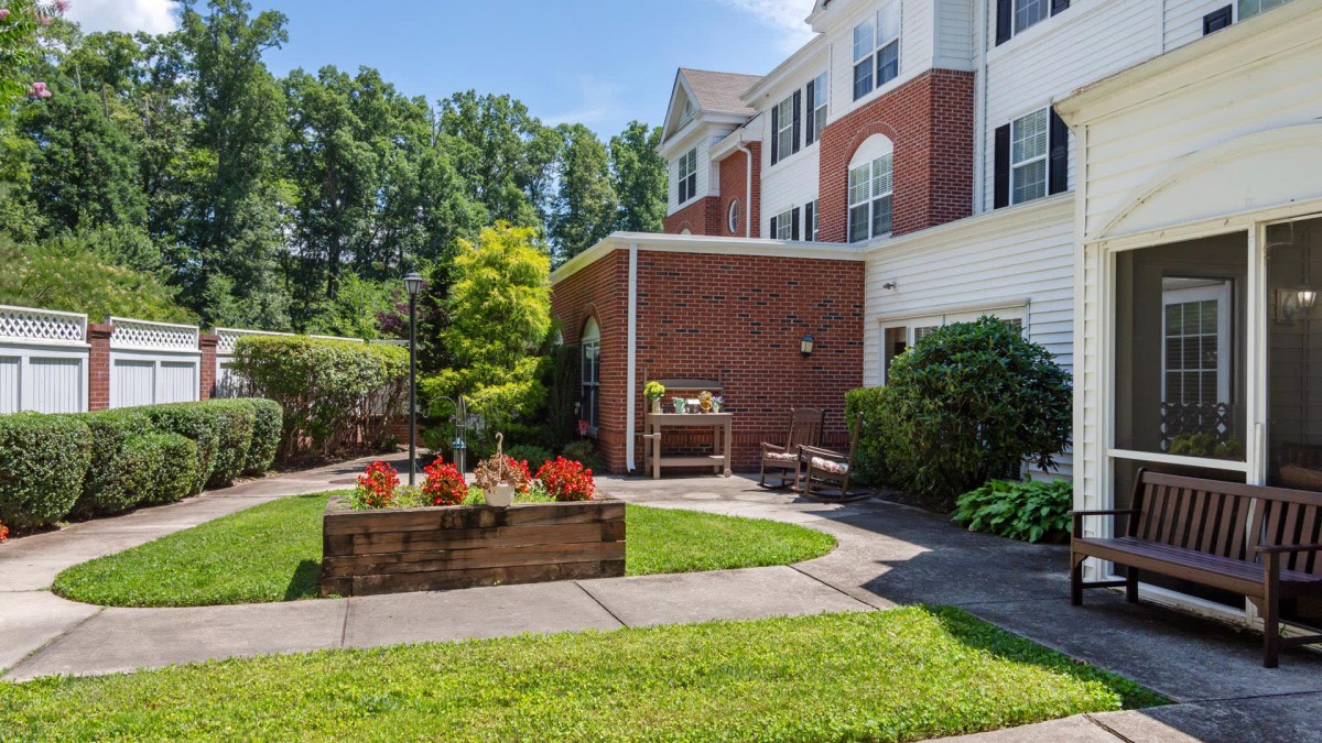 A well-maintained garden area with a brick building in the background, featuring vibrant plants and a grassy lawn.