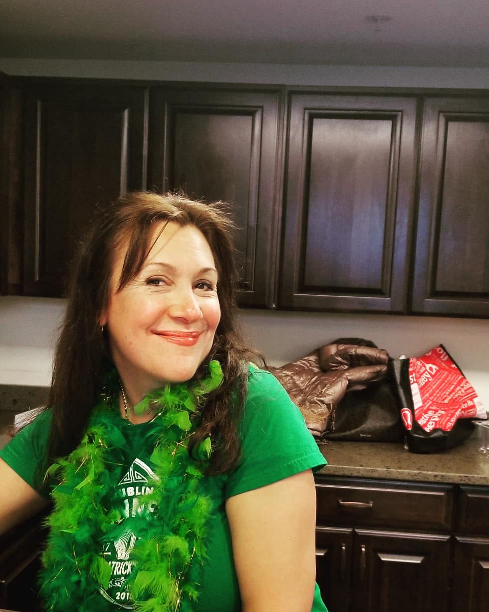 A woman smiling in a kitchen, wearing a green shirt.