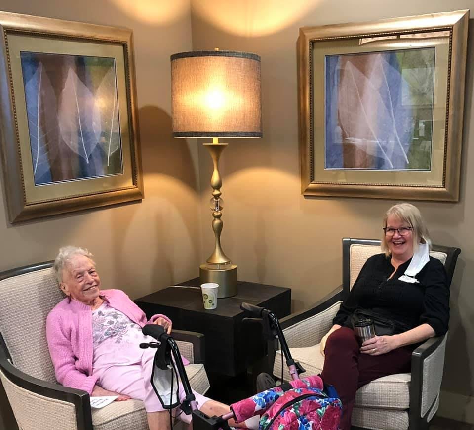 Two elderly women seated and talking in a cozy living room setting with warm lighting.