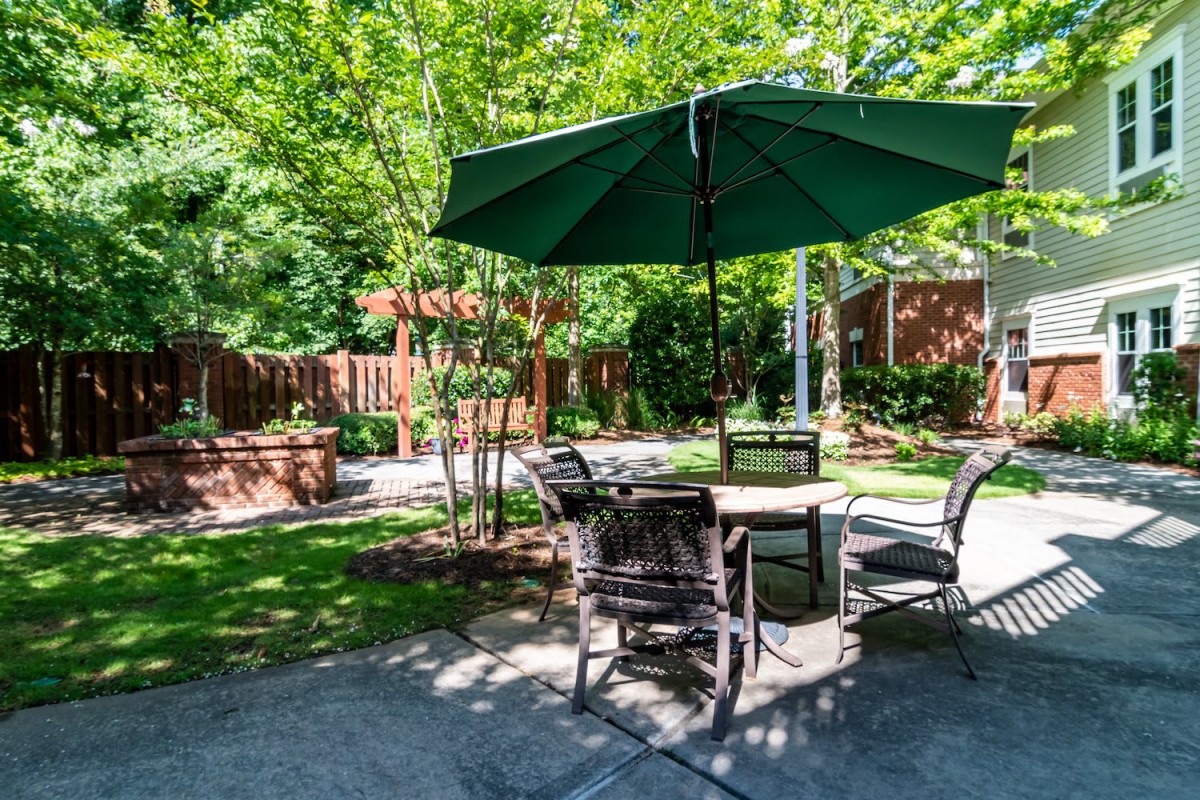 A sunny patio with a table, chairs, and a large green umbrella, surrounded by trees.