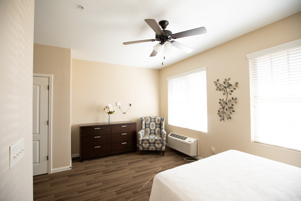 A minimalist bedroom featuring warm colors, a ceiling fan, armchair, and dresser.