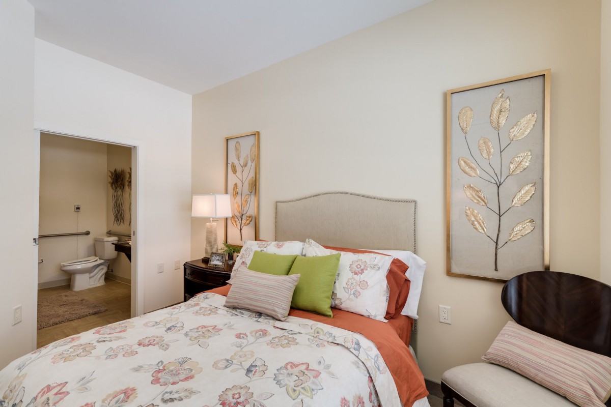 A cozy and beautifully decorated bedroom with a floral comforter, decorative pillows, artwork on the walls, and a bedside lamp.